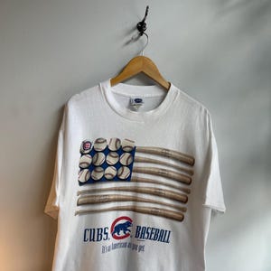 May include: White t-shirt featuring a baseball-themed design. The graphic depicts a baseball bat and ball flag with the text "CUBS BASEBALL" and the phrase "It's as American as you get."