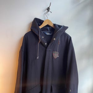 May include: A navy blue hooded coat with a front button closure and two front pockets. The coat features a small emblem on the chest and is hanging on a wooden hanger. The coat has a drawstring hood and is made of a thick material.