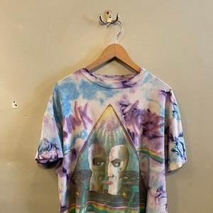 May include: A tie-dye t-shirt featuring a graphic of Pink Floyd's "The Division Bell" album cover. The shirt is white with blue, purple, and pink tie-dye patterns. The album cover features a graphic of two faces with a cityscape in the background. The text "The Division Bell Tour 1994" is printed below the graphic.