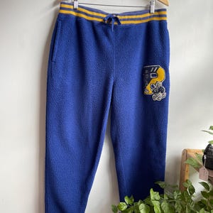 Op de afbeelding: Koningsblauwe fleece joggingbroek met een geel en blauw gestreepte elastische tailleband met trekkoord. De linkerpijp heeft een grote gele patch met de tekst POLO en een cartoonfiguur. De boorden bij de enkels zijn geel en blauw gestreept.