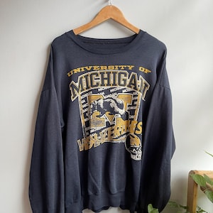 Peut inclure: Un sweat-shirt à col rond gris anthracite foncé avec le texte University of Michigan Wolverines en jaune et blanc. Le motif comprend une illustration de carcajou, un casque de football américain et le mot Michigan répété en arrière-plan.
