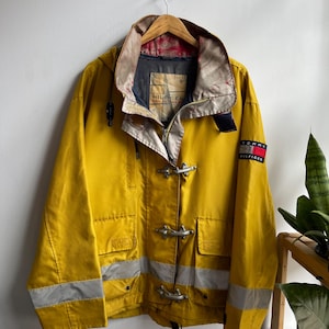 May include: Yellow Tommy Hilfiger sailing jacket with a high collar and toggle closures. The jacket features a reflective stripe on the sleeves and pockets, and a Tommy Hilfiger logo patch. The jacket is hanging on a wooden hanger.
