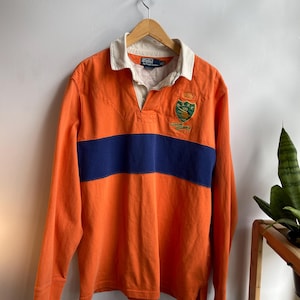 Op de afbeelding: Oranje rugby shirt met lange mouwen en een marineblauwe horizontale streep over de borst. Het shirt heeft een witte kraag en een embleem op de borst. Het shirt hangt aan een houten hanger.
