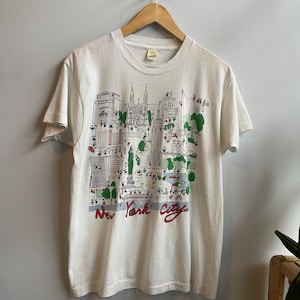 May include: White t-shirt featuring a colorful illustration of New York City landmarks. The design includes buildings, trees, and figures, with "New York City" written in red script across the bottom. The shirt is hanging on a wooden hanger.