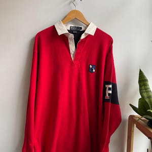 Puede incluir: Una camiseta de rugby roja de manga larga con cuello blanco y un pequeño emblema en el pecho. La manga tiene un panel negro y azul marino con la letra "F" y la palabra "POLO". La camiseta está colgada en una percha de madera.