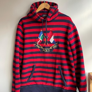 Puede incluir: Sudadera con capucha a rayas rojas y azul marino con bolsillo de canguro. La sudadera tiene un emblema bordado con banderas y la palabra "POLO" y los años 1967 y 1987. La capucha está forrada en azul marino.