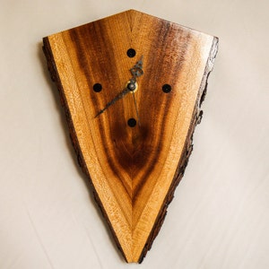 May include: A triangular wooden wall clock with a natural edge. The clock face features a rich, warm wood grain pattern with a dark brown center. Black dots mark the hours, and the clock hands are black.