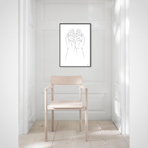 Puede incluir: Una impresi&oacute;n art&iacute;stica minimalista en blanco y negro que presenta un dibujo lineal de dos manos abiertas, enmarcado en negro. La obra de arte cuelga de una pared blanca en una habitaci&oacute;n luminosa, con una silla de madera clara en primer plano. Tema: moderno y sencillo.