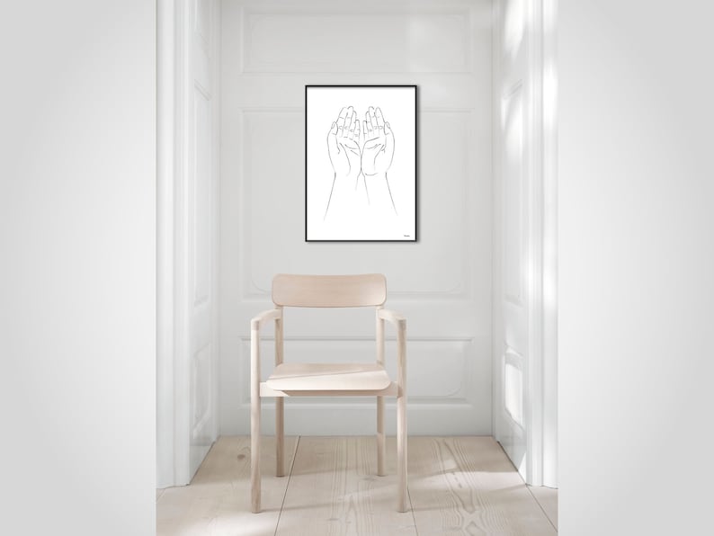 Puede incluir: Una impresi&oacute;n art&iacute;stica minimalista en blanco y negro que presenta un dibujo lineal de dos manos abiertas, enmarcado en negro. La obra de arte cuelga de una pared blanca en una habitaci&oacute;n luminosa, con una silla de madera clara en primer plano. Tema: moderno y sencillo.