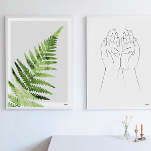 Puede incluir: Dos impresiones art&iacute;sticas enmarcadas. Una presenta un helecho en acuarela en tonos verdes sobre un fondo gris. La otra es un dibujo lineal de dos manos abiertas sobre un fondo blanco. Ambas est&aacute;n en marcos blancos.