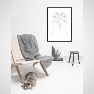 Puede incluir: Una escena interior minimalista con una impresi&oacute;n enmarcada en blanco y negro de manos, una silla tejida, una manta gris y un peque&ntilde;o taburete negro. Un gato gris est&aacute; en el suelo. La habitaci&oacute;n tiene paredes blancas y suelo de madera clara.