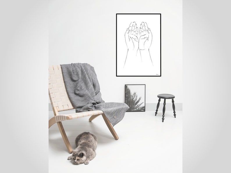 Puede incluir: Una escena interior minimalista con una impresi&oacute;n enmarcada en blanco y negro de manos, una silla tejida, una manta gris y un peque&ntilde;o taburete negro. Un gato gris est&aacute; en el suelo. La habitaci&oacute;n tiene paredes blancas y suelo de madera clara.