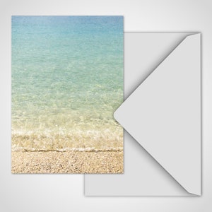 May include: A greeting card featuring a serene beach scene with clear turquoise water and a sandy shore. The card is paired with a white envelope, creating a calming and natural aesthetic. Ideal for sending a message of peace.
