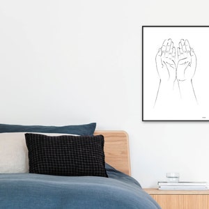Puede incluir: Una impresi&oacute;n enmarcada en blanco y negro con dos manos abiertas, con las palmas hacia arriba. La obra de arte minimalista es adecuada para la decoraci&oacute;n del hogar. La impresi&oacute;n se muestra en una pared blanca sobre una cama con ropa de cama y almohadas azules.