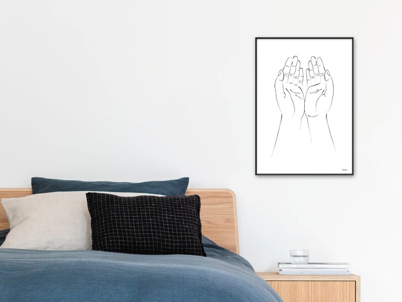 Puede incluir: Una impresi&oacute;n enmarcada en blanco y negro con dos manos abiertas, con las palmas hacia arriba. La obra de arte minimalista es adecuada para la decoraci&oacute;n del hogar. La impresi&oacute;n se muestra en una pared blanca sobre una cama con ropa de cama y almohadas azules.