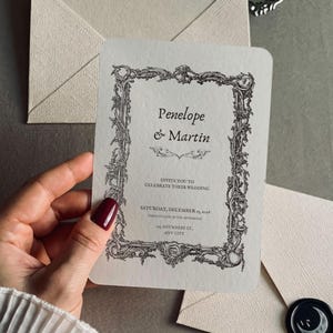 May include: A cream-colored wedding invitation card with rounded corners, featuring a black ornate baroque-style frame. The text reads Penelope & Martin, invite you to celebrate their wedding, Saturday, December 19, 2026, 441 Anywhere St., Any City.