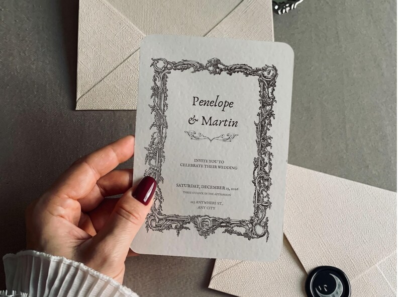 May include: A cream-colored wedding invitation card with rounded corners, featuring a black ornate baroque-style frame. The text reads Penelope & Martin, invite you to celebrate their wedding, Saturday, December 19, 2026, 441 Anywhere St., Any City.