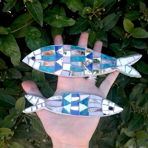 Può includere: Due decorazioni da parete a forma di pesce in mosaico, una blu e bianca, l'altra argentata e blu, con una mano che le tiene.