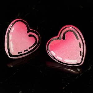 Puede incluir: Dos pendientes en forma de corazón rosa con contornos negros. Los corazones tienen un acabado brillante.