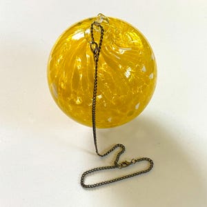 May include: A round, yellow glass ornament with a swirled pattern and white accents. A dark metal chain is attached to the top and hangs down. The ornament is a decorative item.