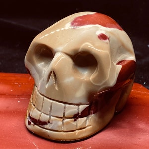 May include: A carved skull made of red, white, and brown jasper. The skull has a detailed face with a wide smile and a prominent jawline.