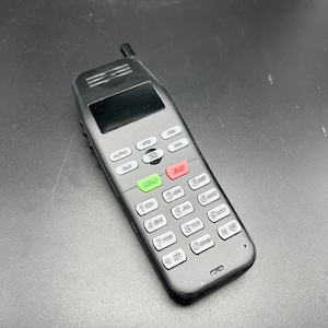 Puede incluir: Un teléfono negro con cable, con una pequeña pantalla y un teclado con botones blancos. Los botones tienen letras y números. El teléfono tiene una pequeña antena en la parte superior.