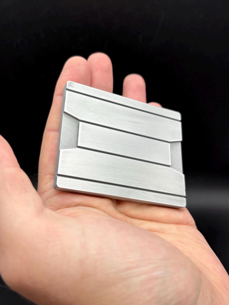 Pode incluir: Um objeto retangular de metal escovado com linhas horizontais e detalhes geom&eacute;tricos. O objeto &eacute; segurado em uma m&atilde;o contra um fundo preto. O metal tem uma cor prateada.