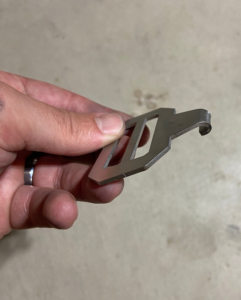 May include: A silver metal hook with a rectangular loop. The hook is designed to be attached to a belt or bag.