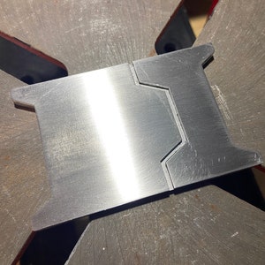 May include: A close-up of a silver metal piece with a smooth, brushed finish. The piece has a unique shape with two cutouts on one side. The piece is likely a part of a larger metal object.