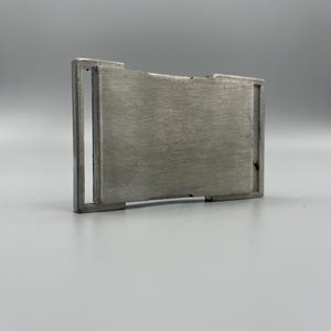 May include: A brushed metal belt buckle with a rectangular design. The buckle has a central plate and a surrounding frame. The metal has a matte finish and a silver color. The buckle is designed to be used with a belt.