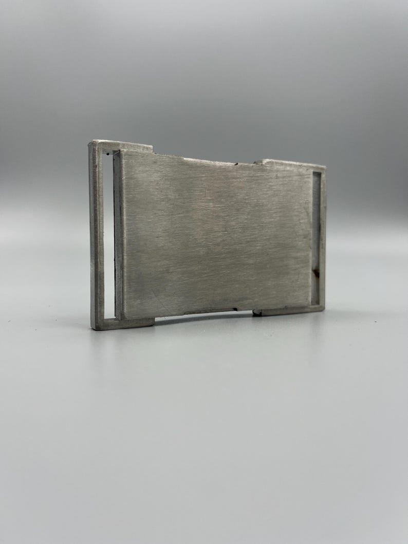 May include: A brushed metal belt buckle with a rectangular design. The buckle has a central plate and a surrounding frame. The metal has a matte finish and a silver color. The buckle is designed to be used with a belt.