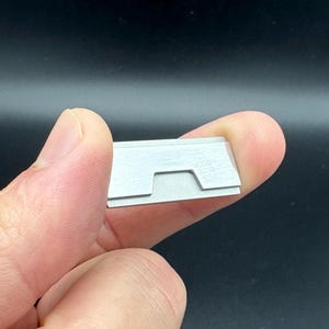 Op de afbeelding: Een klein, rechthoekig, zilverkleurig metalen object met een inkeping. Het object wordt tussen twee vingers tegen een donkere achtergrond gehouden. Het lijkt een klein gereedschap of onderdeel te zijn.