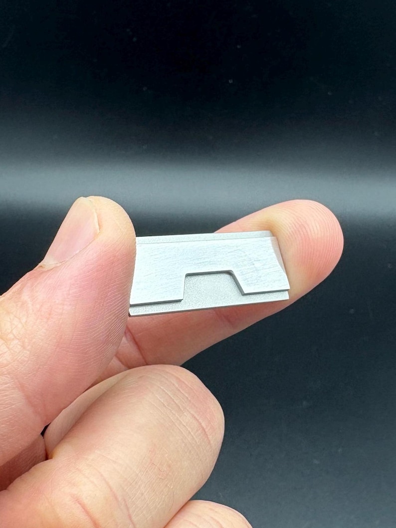 Op de afbeelding: Een klein, rechthoekig, zilverkleurig metalen object met een inkeping. Het object wordt tussen twee vingers tegen een donkere achtergrond gehouden. Het lijkt een klein gereedschap of onderdeel te zijn.