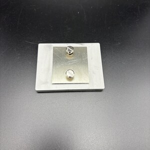 Pode incluir: Uma pequena placa de metal retangular com uma placa de metal quadrada menor fixada no topo. Dois parafusos prateados s&atilde;o vis&iacute;veis, um perto do topo e outro perto da parte inferior da placa menor. O fundo &eacute; de cor cinza escuro.
