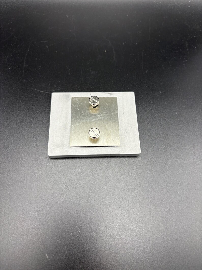 Pode incluir: Uma pequena placa de metal retangular com uma placa de metal quadrada menor fixada no topo. Dois parafusos prateados s&atilde;o vis&iacute;veis, um perto do topo e outro perto da parte inferior da placa menor. O fundo &eacute; de cor cinza escuro.