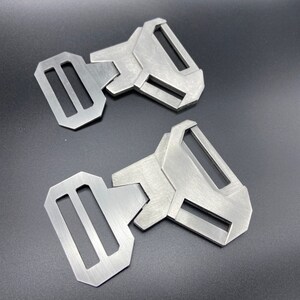 May include: Two silver metal belt buckles with a geometric design. The buckles have a rectangular shape with a rounded edge and a single, long, rectangular slot.