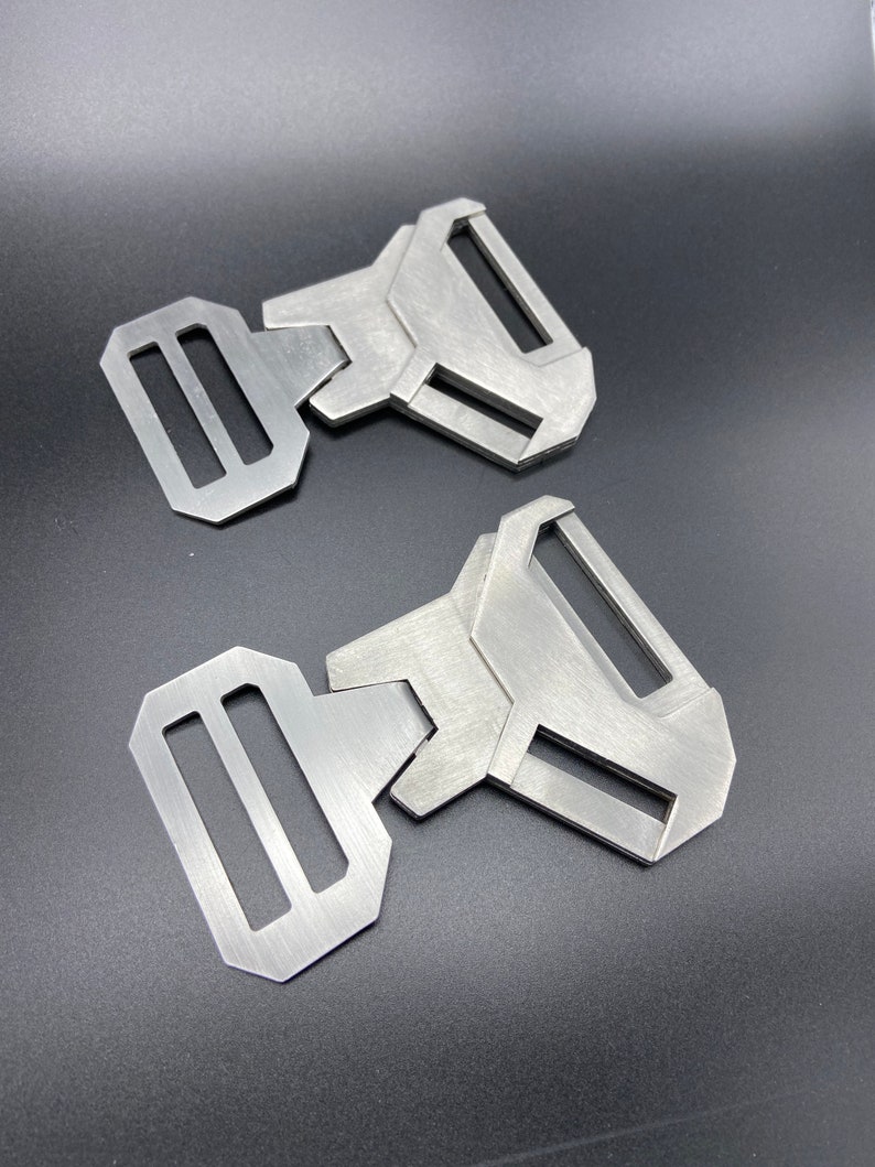 May include: Two silver metal belt buckles with a geometric design. The buckles have a rectangular shape with a rounded edge and a single, long, rectangular slot.