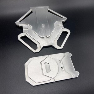 May include: Two silver metal pieces, one shaped like a star with a hole in the centre, and the other a rectangular shape with a rounded edge and a hole in the centre.
