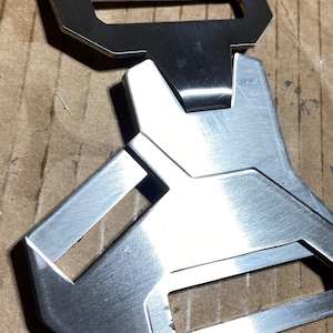 May include: A silver metal buckle with a rectangular shape and a unique design. The buckle has two openings, one on the top and one on the bottom.