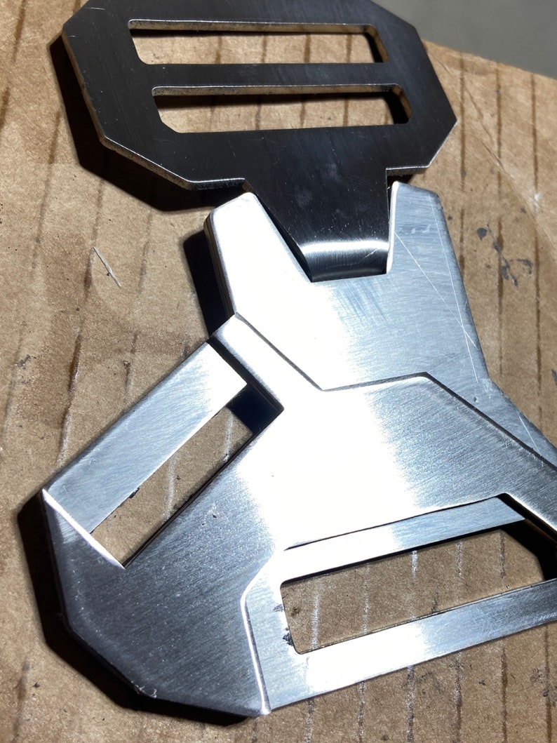 May include: A silver metal buckle with a rectangular shape and a unique design. The buckle has two openings, one on the top and one on the bottom.