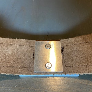 May include: A close-up of a metal plate with two screws attached to a piece of brown leather.