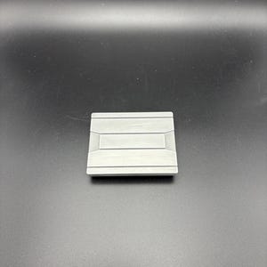 Pode incluir: Um objeto met&aacute;lico quadrado prateado com um design retangular rebaixado no centro. O objeto tem uma superf&iacute;cie ligeiramente texturizada e parece ser uma pequena pe&ccedil;a decorativa ou um componente de hardware.