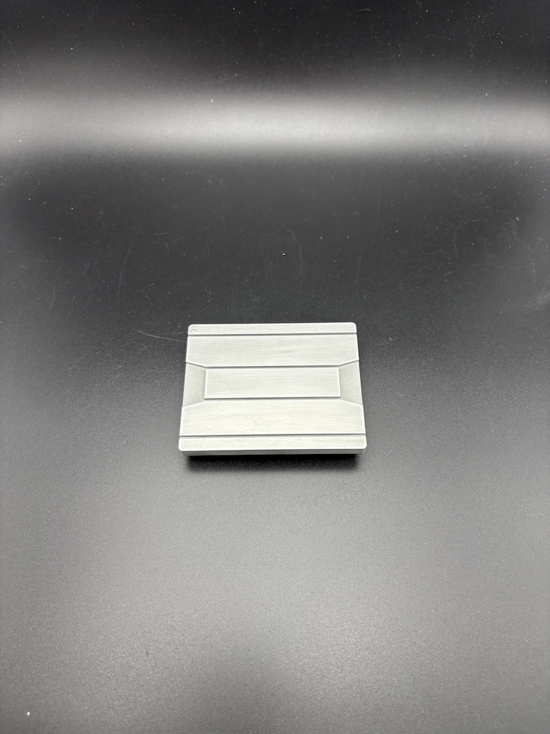 Pode incluir: Um objeto met&aacute;lico quadrado prateado com um design retangular rebaixado no centro. O objeto tem uma superf&iacute;cie ligeiramente texturizada e parece ser uma pequena pe&ccedil;a decorativa ou um componente de hardware.