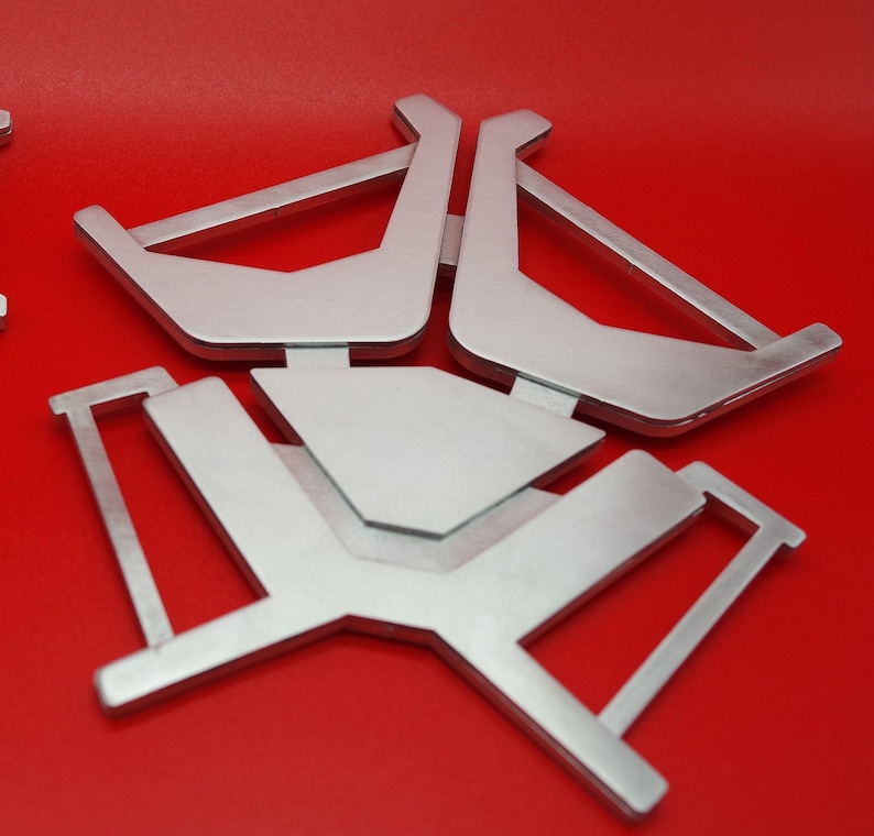May include: Silver metal stand with three legs and a rectangular top. The stand is designed to hold a small object, such as a phone or a tablet.