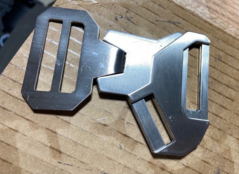 May include: A silver metal buckle with a rectangular shape and two slots. The buckle has a brushed finish and a geometric design.