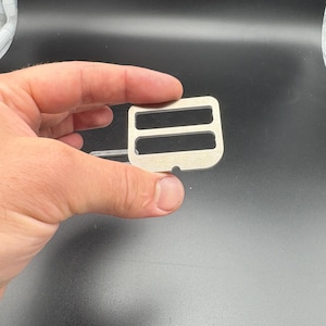 Puede incluir: Una hebilla met&aacute;lica de color plateado con dos ranuras rectangulares y un peque&ntilde;o agujero. La hebilla se sostiene en una mano sobre un fondo oscuro. Este componente de hardware es probablemente para una bolsa o correa.