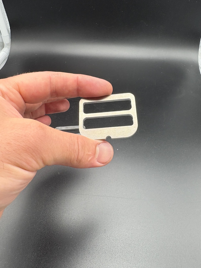 Puede incluir: Una hebilla met&aacute;lica de color plateado con dos ranuras rectangulares y un peque&ntilde;o agujero. La hebilla se sostiene en una mano sobre un fondo oscuro. Este componente de hardware es probablemente para una bolsa o correa.