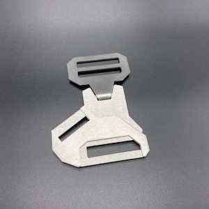 May include: A silver metal buckle with two slots for a strap. The buckle has a unique design with a triangular shape and a smooth, brushed finish.