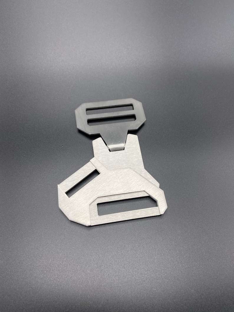 May include: A silver metal buckle with two slots for a strap. The buckle has a unique design with a triangular shape and a smooth, brushed finish.