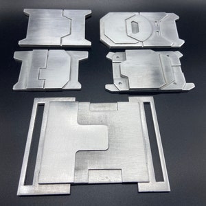 K&ouml;nnte beinhalten: F&uuml;nf Metallteile, m&ouml;glicherweise f&uuml;r ein DIY-Projekt, sind auf einer schwarzen Oberfl&auml;che angeordnet. Die Teile sind silberfarben und haben eine geb&uuml;rstete Oberfl&auml;che. Das gr&ouml;&szlig;te Teil ist rechteckig mit einem Ausschnitt in der Mitte. Die anderen Teile sind kleiner und haben verschiedene Formen.